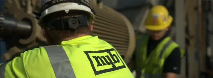 Two construction workers in neon safety vests and hard hats in an industrial facility, one foreground with a logo on the vest.