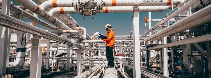Industrial oil and gas facility with a worker in orange safety gear inspecting white pipes.