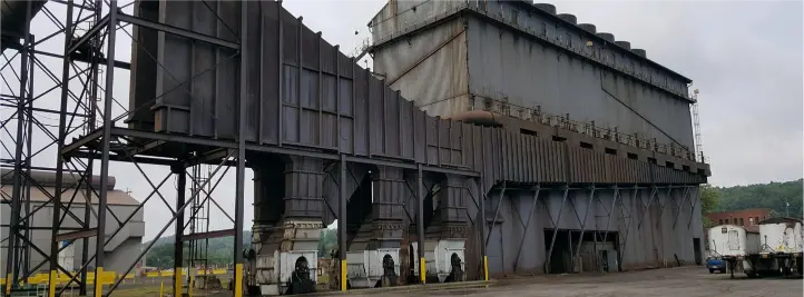 Large gray industrial facility with a raised conveyor structure, heavy metal supports, and workers standing near the loading area.