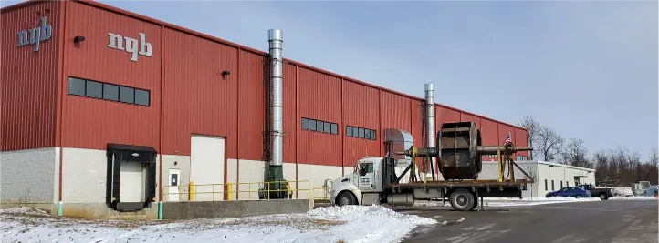 Red industrial warehouse with loading dock; a flatbed truck carries a large spool on a trailer in a snowy lot.