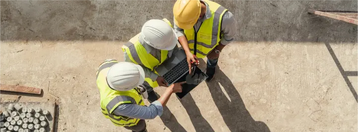 Three construction workers in hard hats and high-visibility vests examine a laptop on a dirt construction site, collaborating over a tablet-like device.