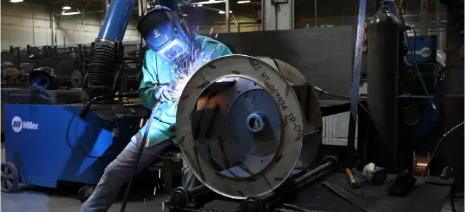 Welder wearing a protective helmet and blue gear, welding a large circular metal flange in a factory setting.