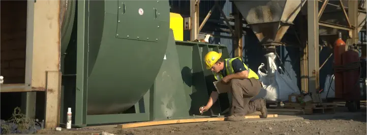 Maintenance worker in a yellow helmet crouches beside a large green industrial unit outdoors, inspecting a panel edge.