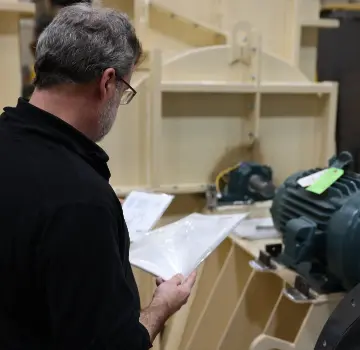 Man with gray hair and glasses inspecting papers at a woodworking shop, standing beside a large machine.