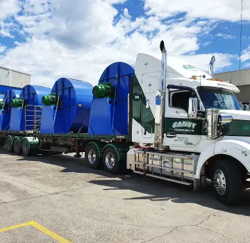 White semi-truck with trailer carrying several large blue cylindrical wind turbine components secured with straps in an industrial yard.