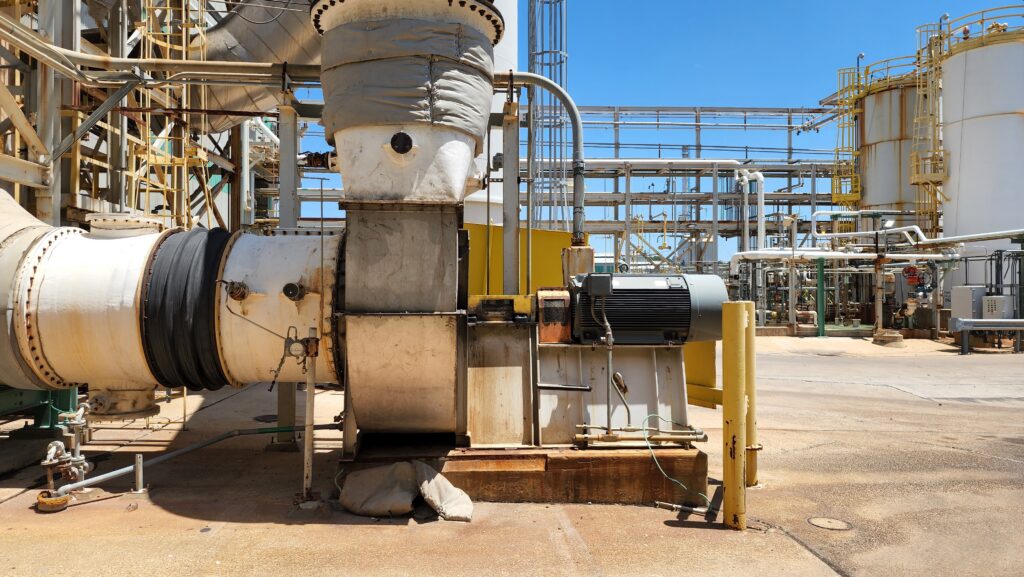Industrial processing site with large metal pipes, ducts, and tanks outdoors under a clear blue sky.